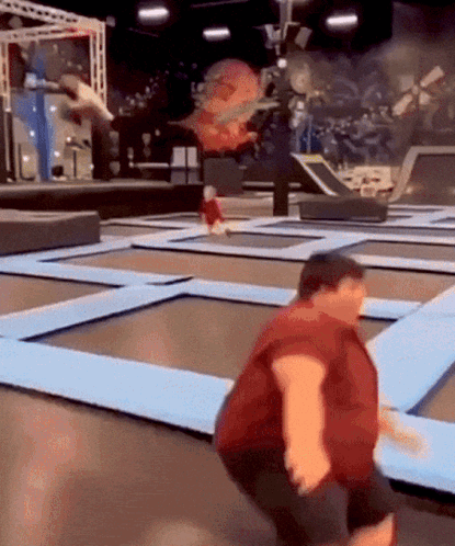 A Man In A Red Shirt Is Jumping On A Trampoline With A Child In The Background . GIF