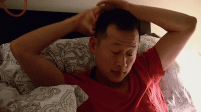 A Man In A Red Shirt Is Laying In Bed With His Hands On His Head GIF