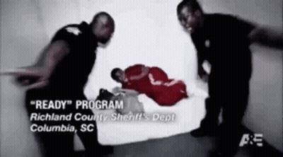 A Man In A Red Shirt Is Laying On The Floor While Two Police Officers Stand Around Him . GIF