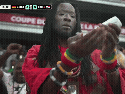 A Man In A Red Shirt Is Looking At A Cell Phone In Front Of A Scoreboard That Says Gha 0 Pan GIF