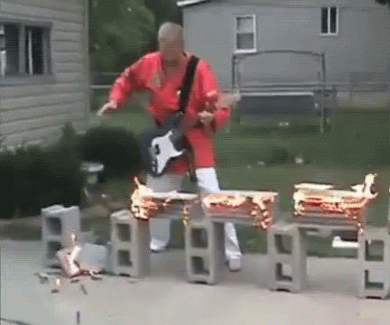 A Man In A Red Shirt Is Playing A Guitar In Front Of Some Burning Drums GIF