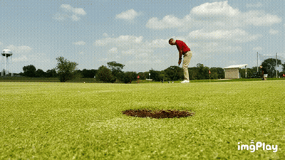 A Man In A Red Shirt Is Putting A Ball Into A Hole On A Golf Course GIF