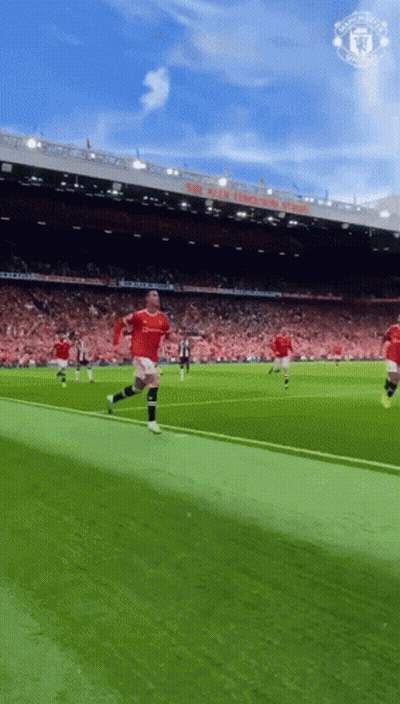 A Man In A Red Shirt Is Running On A Soccer Field In Front Of A Manchester United Stadium GIF