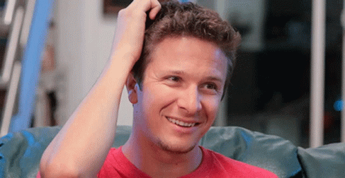 A Man In A Red Shirt Is Scratching His Head And Smiling GIF