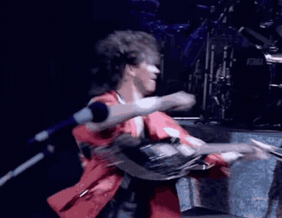 A Man In A Red Shirt Is Singing Into A Microphone In Front Of A Tama Drum Set GIF