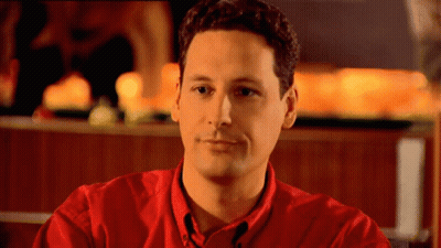 A Man In A Red Shirt Is Sitting In Front Of A Fireplace GIF