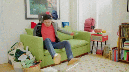 A Man In A Red Shirt Is Sitting On A Green Couch In A Messy Living Room . GIF