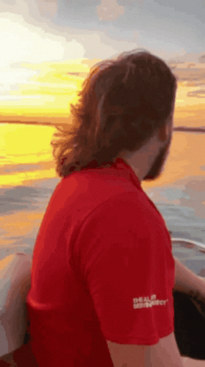 A Man In A Red Shirt Is Sitting On The Back Of A Boat Looking At The Sunset . GIF