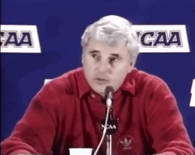 A Man In A Red Shirt Is Speaking Into A Microphone With Ncaa Logos In The Background GIF