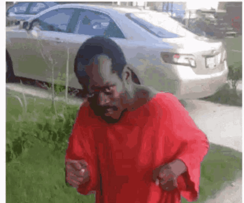 A Man In A Red Shirt Is Standing In Front Of A Silver Car . GIF