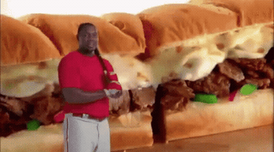 A Man In A Red Shirt Is Standing In Front Of A Very Large Sandwich . GIF