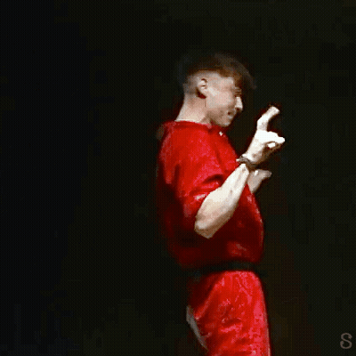 A Man In A Red Shirt Is Standing On A Stage With His Hands Up GIF