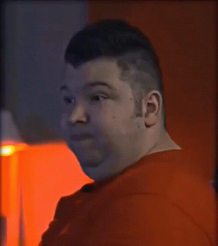 A Man In A Red Shirt Looks At The Camera In A Dark Room GIF
