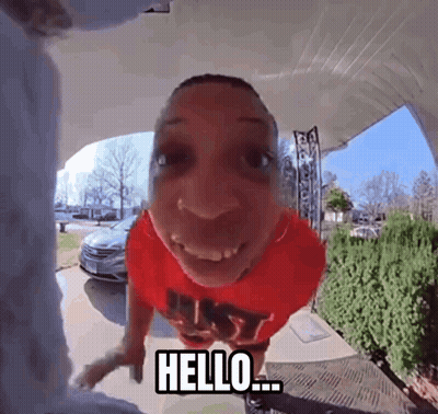 A Man In A Red Shirt Says Hello While Standing In Front Of A Door GIF