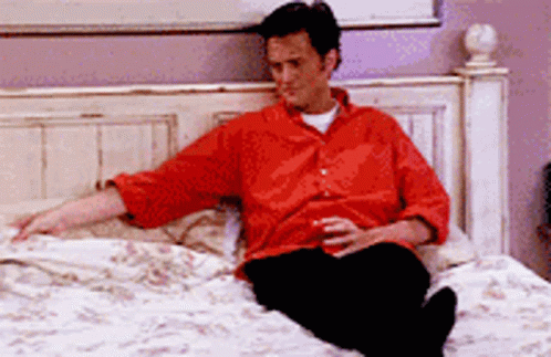 A Man In A Red Shirt Sits On A Bed GIF