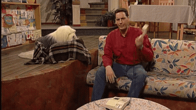 A Man In A Red Shirt Sits On A Couch With A Dog Behind Him GIF