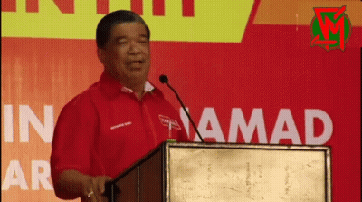 A Man In A Red Shirt Stands At A Podium In Front Of A Sign That Says In Amada GIF