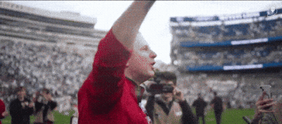 A Man In A Red Shirt Stands In A Stadium With His Arms In The Air GIF