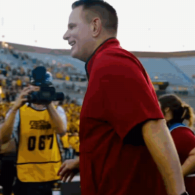 A Man In A Red Shirt Stands In Front Of A Camera Wearing A Yellow Vest With The Number 067 On It GIF