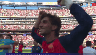 A Man In A Red Shirt Stands In Front Of A Stadium With A Sign That Says The U GIF