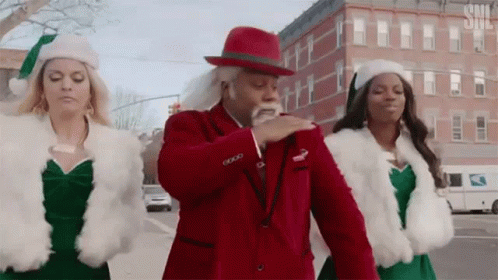 A Man In A Red Suit And White Hat Is Dancing With Two Women In Santa Hats . GIF