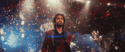 A Man In A Red Suit Stands On A Stage With Confetti Falling Around Him And A Sign That Says Peace Makes Love Stronger GIF