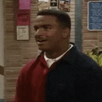 A Man In A Red Sweater And Black Jacket Is Standing In Front Of A Brick Wall . GIF