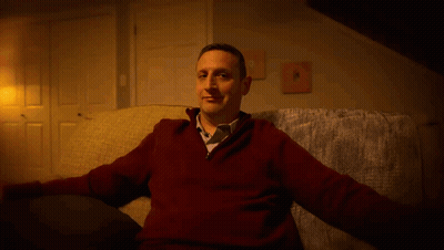 A Man In A Red Sweater And Tie Is Sitting On A Couch GIF