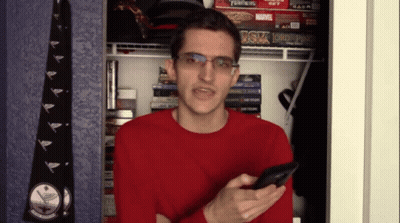 A Man In A Red Sweater Is Holding A Cell Phone In Front Of A Shelf With A Board Game That Says Transformers GIF