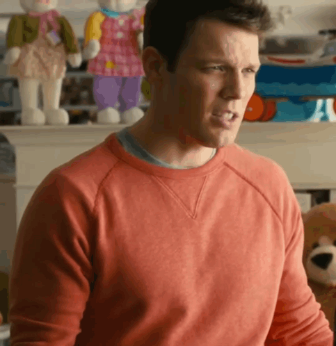 A Man In A Red Sweater Is Sitting In Front Of A Fireplace With Stuffed Animals On It GIF