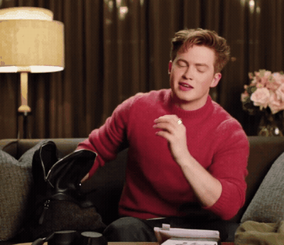 A Man In A Red Sweater Sits On A Couch Holding A Black Bag GIF