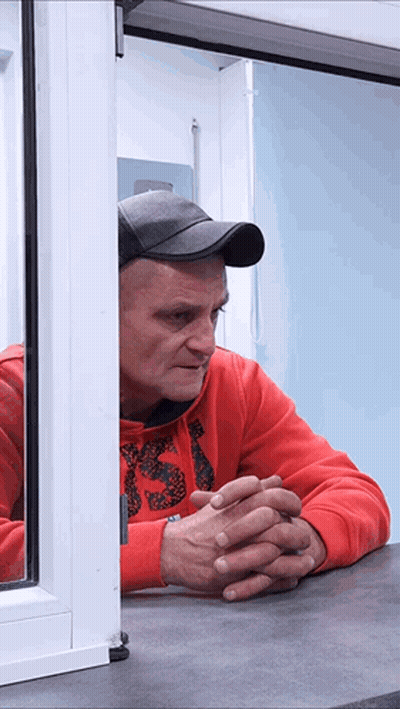 A Man In A Red Sweatshirt With The Letter S On It Looks Out Of A Window GIF