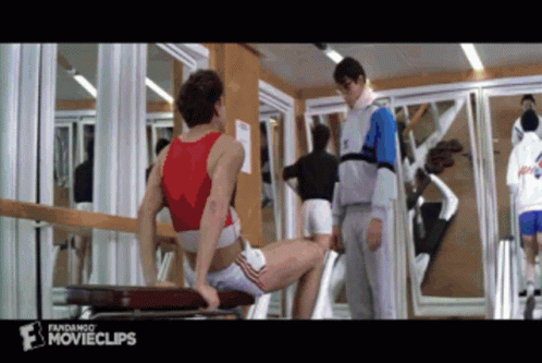 A Man In A Red Tank Top Sits On A Bench In A Gym While A Man In A Grey Jacket Stands Behind Him GIF