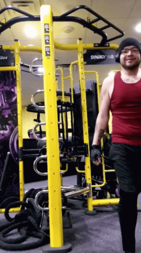 A Man In A Red Tank Top Walks In Front Of A Gym Machine That Says Synergy 360 GIF