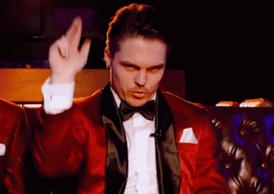 A Man In A Red Tuxedo And Bow Tie Is Sitting On A Couch Making A Gesture With His Hand . GIF