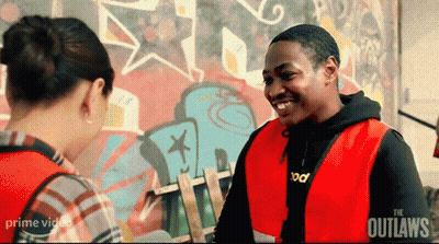A Man In A Red Vest Is Smiling While Talking To A Woman In Front Of A Graffiti Wall . GIF