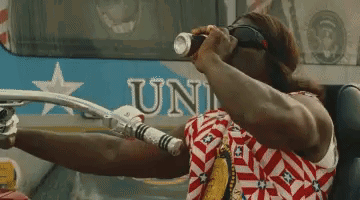 A Man In A Red White And Blue Shirt Is Drinking From A Can While Sitting In Front Of A Bus That Says Uni GIF
