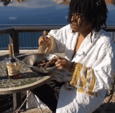 A Man In A Robe Is Sitting At A Table With A Bottle Of Hennessy GIF
