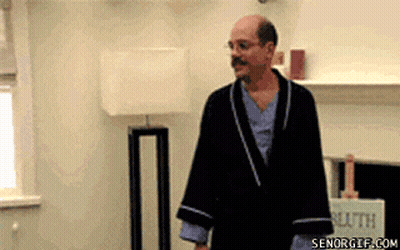 A Man In A Robe Is Standing In A Living Room With A Lamp . GIF