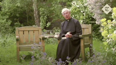 A Man In A Robe Sits In A Chair In A Garden With A T On The Corner GIF
