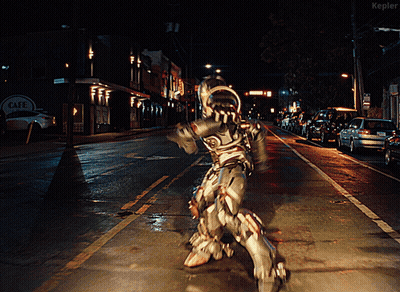A Man In A Robot Costume Is Walking Down A Street At Night In Front Of A Cafe Sign GIF