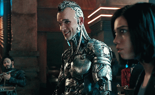 A Man In A Robotic Suit Stands Next To A Woman In A Dark Room GIF