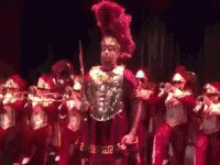 A Man In A Roman Costume Is Standing In Front Of A Marching Band Holding A Sword . GIF