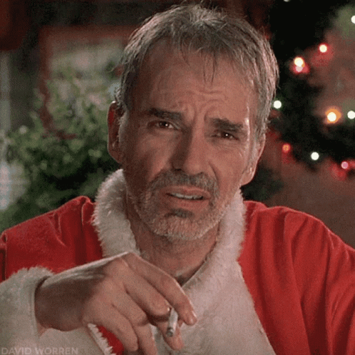 A Man In A Santa Claus Outfit Is Smoking A Cigarette In Front Of A Christmas Tree GIF