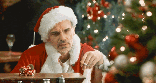 A Man In A Santa Suit Is Smoking A Cigarette In Front Of A Christmas Tree GIF