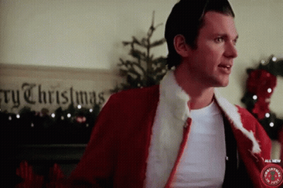 A Man In A Santa Suit Is Standing In Front Of A Sign That Says " Merry Christmas " GIF