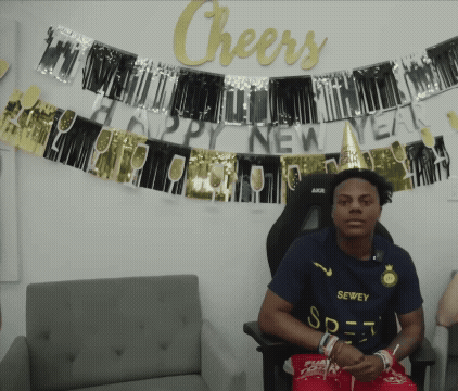 A Man In A Sewee Shirt Sits In Front Of A Sign That Says Cheers GIF