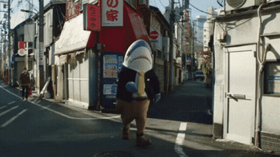 A Man In A Shark Costume Walks Down A Street In Front Of A Store With Chinese Writing On It GIF