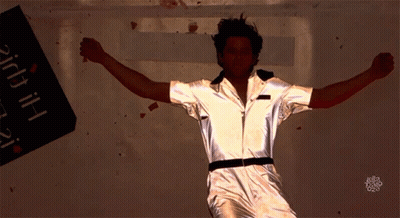 A Man In A Silver Jumpsuit Is Dancing In Front Of A Sign That Says ' Bitch ' GIF
