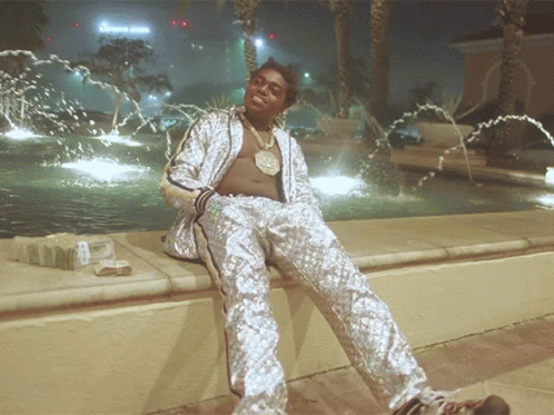 A Man In A Silver Suit Sits In Front Of A Fountain With A Sign That Says Green Juice GIF